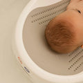 A baby with wet hair lies partially submerged in water on a white Nuby UK Newborn Bath Support & Thermometer, showing the back of the babys head and the upper part of the bath support.