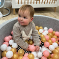 A baby with rosy cheeks sits in the Nuby Ball Pit for Babies, surrounded by pink, gold, and white balls, wearing a beige sweater with a white bunny. A crib appears in the background.