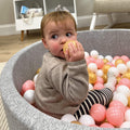 A toddler enjoys the Nuby Ball Pit for Babies, filled with pink, white, and gold balls. The soft pit, set in a cozy room with light furniture, creates a gentle play space perfect for little ones.