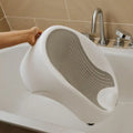 A hand places the Nuby UK Newborn Bath Support & Thermometer, featuring a cushioned, perforated gray surface, into a bathtub beside a silver faucet and controls.