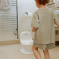 A young child with short curly hair stands in a bathroom, facing the Nuby UK Potty Training Kit Bundle placed beside a towel rack, toilet paper holder, and sink.