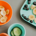 An overhead view of the Brights Complete Weaning Set by Nuby UK: an orange bowl with sliced bananas, a teal cup with cucumber slices, and a teal plate holding blueberries and banana slices—ideal weaning essentials for little ones.