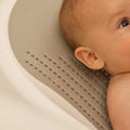 A close-up of the Nuby UK Newborn Bath Support & Thermometer in use, showing a baby lying in a light gray tub with water droplets on the perforated surface, focusing on the babys head, ear, and shoulder.