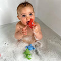 A baby enjoys a bubble bath, chewing on a red Nuby UK toy with a face while a green alligator and blue whale from the Nuby UK Baby Bath Sets for Newborns 0-12 Months float nearby—ideal for a baby shower gift or newborn bath set.