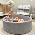 A baby sits in the Nuby Ball Pit for Babies, filled with pastel balls in a nursery featuring a crib, a white shelf with baby items, and a light-colored floor.