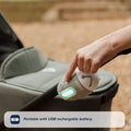 A hand holds a Nuby UK Jitterbug Pram Rocker with a glowing green light, scanning the handle of a stroller outdoors. Text overlay reads: Portable with USB rechargeable battery.