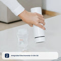 A hand holds a white Nuby RapidCool™ Baby Bottle Maker above a baby bottle on a table. Text below reads, Lid features an integrated thermometer—ideal for travel feeding with the RapidCool™ twin pack.