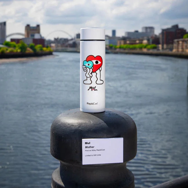 A white limited edition RapidCool x Mul baby bottle by Nuby, featuring a colorful cartoon heart design, stands on a riverside railing with cityscape and river in the background. The “Mul Mother” label highlights this exclusive release.