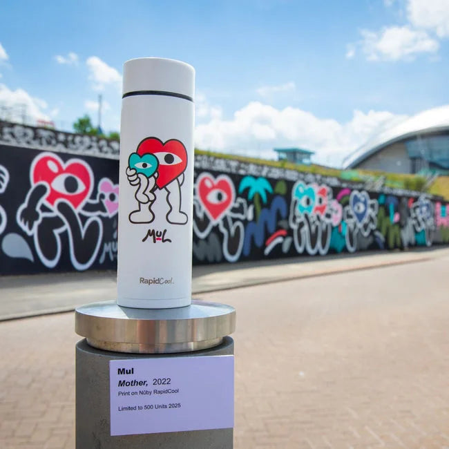 A white Nuby RapidCool x Mul bottle with vibrant cartoon heart art stands on a pedestal outdoors, with a mural of colorful hearts behind it under a partly cloudy sky.