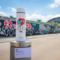 A white Nuby RapidCool x Mul bottle with vibrant cartoon heart art stands on a pedestal outdoors, with a mural of colorful hearts behind it under a partly cloudy sky.