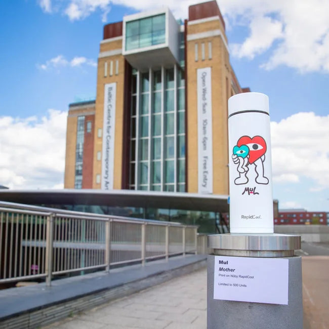 The limited edition Nuby RapidCool x Mul baby bottle, featuring colorful cartoon art, stands on a ledge outside a modern brick building with large glass windows and vertical banners under a partly cloudy sky.