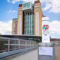 The limited edition Nuby RapidCool x Mul baby bottle, featuring colorful cartoon art, stands on a ledge outside a modern brick building with large glass windows and vertical banners under a partly cloudy sky.