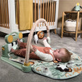 A baby lies on the Nuby Animal Adventures Piano Play Mat & Gym, reaching for sensory hanging toys with their feet beside the toy piano. A crib and a basket are visible in the background.