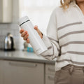 A person in a striped sweater holds the sleek white Nuby UK RapidCool™ Bottle Maker and UV Steriliser Bundle in a modern kitchen, with a kettle visible in the background.