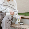 Sitting on a wooden bench outdoors, a person in a white and gray striped sweater and khaki pants pours milk into a baby bottle, possibly prepping for feeding with the Nuby UK RapidCool™ Kit and UV Steriliser Bundle.