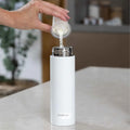 A hand pours powdered formula into a white Nuby RapidCool™ Baby Bottle Maker Twin Pack on a light countertop, with a blurred plant and kitchen items in the background—perfect for travel bottle feeding.