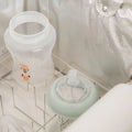 A clear baby bottle with a cartoon animal design and separate nipple sit on a metal rack against a silver background—ideal for formula feeding with the RapidCool™ Bottle Maker and UV Steriliser Bundle by Nuby UK.