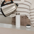 A person pours hot water from a stainless steel kettle into a white Nuby UK RapidCool™ Bottle Maker—ideal for formula feeding—on a kitchen counter, with the bottle maker’s cap placed nearby.
