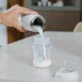 A hand pours milk from a white thermos into a clear baby bottle with a bear design, next to the cap and nipple—perfect to use after UV sterilisation in your Nuby UK RapidSterilise Baby Bottle Maker Steriliser.