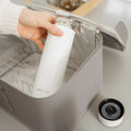A hand places a white bottle from the Nuby UK RapidCool™ Kit and UV Steriliser Bundle into a gray insulated bag, while the circular UV steriliser rests on the table nearby.