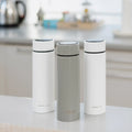 Three upright cylindrical mugs—two white, one gray—resemble the sleek Nuby UK RapidCool™ Kit and UV Steriliser Bundle, with the gray mug in the center flanked by the white ones on a kitchen counter.