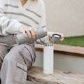 A person in a striped sweater and cargo pants sits on wooden steps, pouring water from a gray bottle into a white insulated one with its lid nearby—possibly preparing to use the Nuby UK RapidCool™ Kit and UV Steriliser Bundle. Grass and a beige wall are behind them.