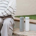 A person in a striped sweater and cargo pants sits on a bench beside two insulated travel mugs and the Nuby UK RapidCool™ Twin Pack and UV Steriliser Bundle, with grass and a concrete wall behind them.