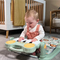 A baby in a white shirt and pink overalls plays on the Nuby Animal Adventures Piano Play Mat & Gym with a green and yellow toy piano, sitting in front of a wooden crib.