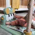 A baby in orange pants enjoys the Nuby Animal Adventures Piano Play Mat & Gym, kicking the toy piano with their feet as sensory hanging toys dangle above. A crib and yellow blanket can be seen in the background.
