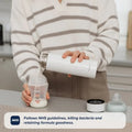 A person in a striped sweater pours liquid from a white Nuby RapidCool x Mul thermos into a baby bottle on a kitchen counter. Text reads: Follows NHS guidelines, kills bacteria, and retains formula goodness.