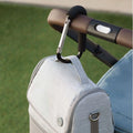 The Nuby UK Pram and Pushchair Accessories Bundle, in light gray, is attached to a strollers brown handle with a black carabiner; the stroller has blue fabric and is set against green grass.