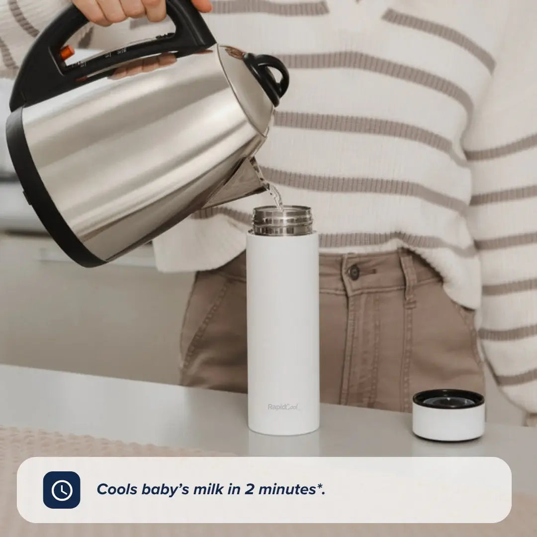 A person pours hot water from an electric kettle into a white Nuby RapidCool x Mul formula feed cooler on a kitchen counter. On-screen text reads, Cools baby’s milk in 2 minutes*.
