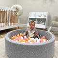 A baby enjoys the Nuby Ball Pit for Babies, filled with pink, white, and gold balls, in a nursery featuring a crib, toy shelves, and a light-colored armchair.