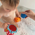 A toddler sits in a bubbly bath, playing with colorful Nuby UK Baby Bath Sets for Newborns 0-12 Months, while an adult pours water from a blue toy cup—an ideal gift for baby showers.