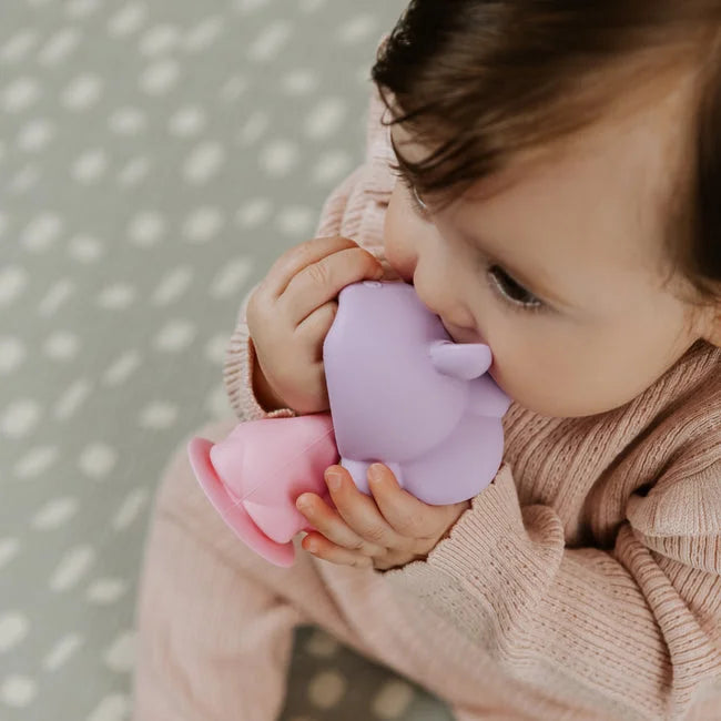 A baby in a pink outfit sits on a patterned mat, holding and chewing on the Nuby Bobble Highchair & Teething Toy, a purple and pink silicone animal-shaped teether.