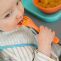 A baby wearing a striped bib eats with an orange spoon-fork and a bowl from the Nuby UK Brights Complete Weaning Set, with more tableware on a teal tray in the background.