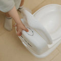 A child, dressed in a light outfit and socks, holds the handle of the Nuby UK Potty Training Kit Bundle on a light-colored floor.