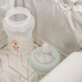 A clear baby bottle with a cartoon animal design and a bottle nipple with a pale green ring sit in a drying rack inside the metallic-lined Nuby 3 Minute UV Baby Steriliser.