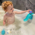 A smiling young child plays in a bubble-filled tub, placing a purple ball from the Nuby UK Bath Toy Bundle 12 Months + at the top of a blue slide attached to the tub wall, while other baby bath toys float nearby.