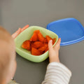 A young child holds a light green bowl from the Nuby UK Brights Complete Weaning Set, filled with orange fruit pieces, on a gray surface. A blue lid rests nearby—ideal for your weaning essentials.